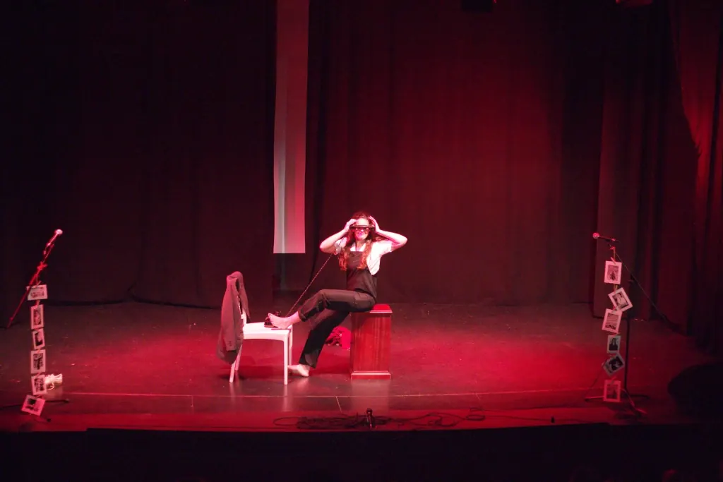 Alba Berrocal encarnando a Dora Maar, sentada sobre el atril rojo con el brazo alzado, bajo luz roja de escenario