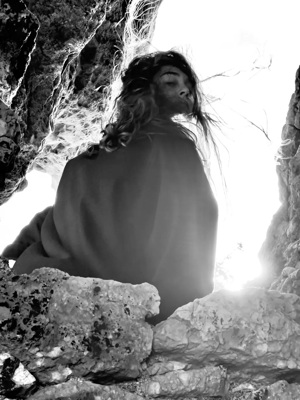 Alba Berrocal de espaldas entre rocas a contraluz, con el pelo al viento, fotografía en blanco y negro
