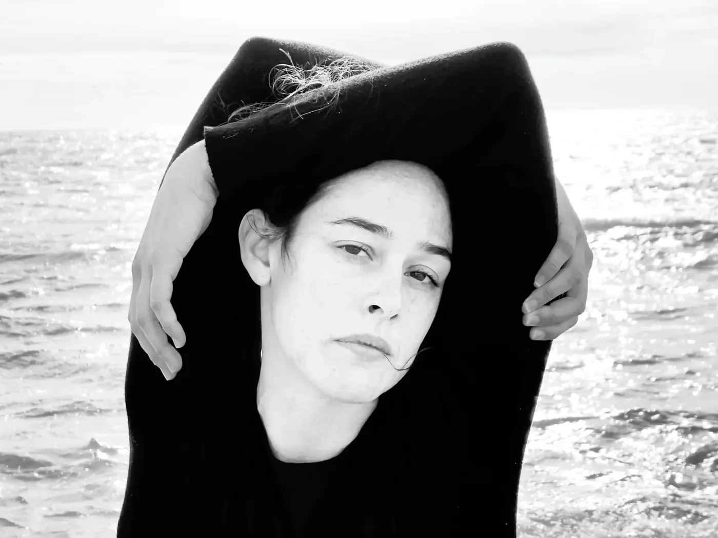 Retrato de Alba Berrocal en primer plano frente al mar, con un jersey oscuro cubriéndole la cabeza